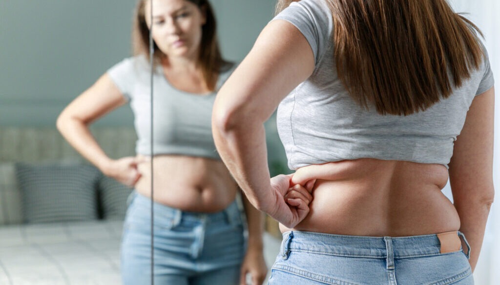 Woman standing in front of mirror looking at the stubborn fat around her waist.